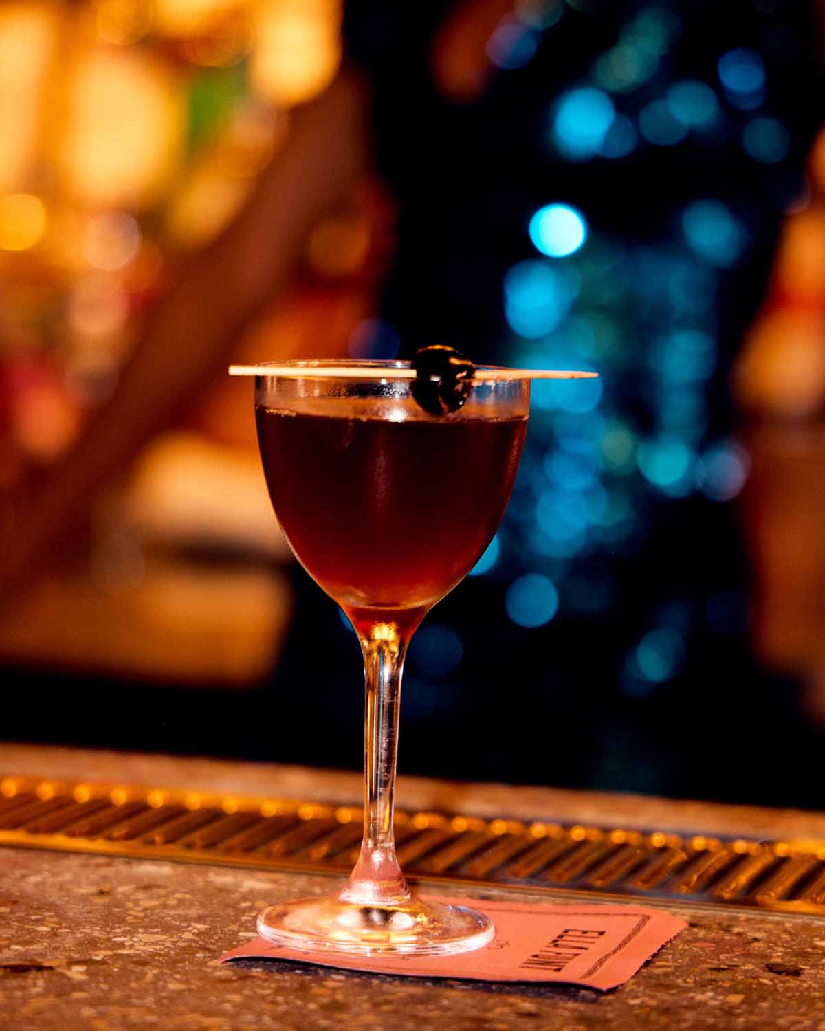 Red cocktail in a glass with a cherry on a bar counter, blurred bar background