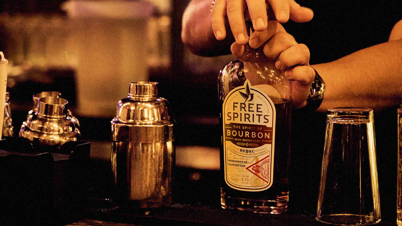 Person pouring Free Spirits bourbon alternative  into a glass with bar equipment in the background.