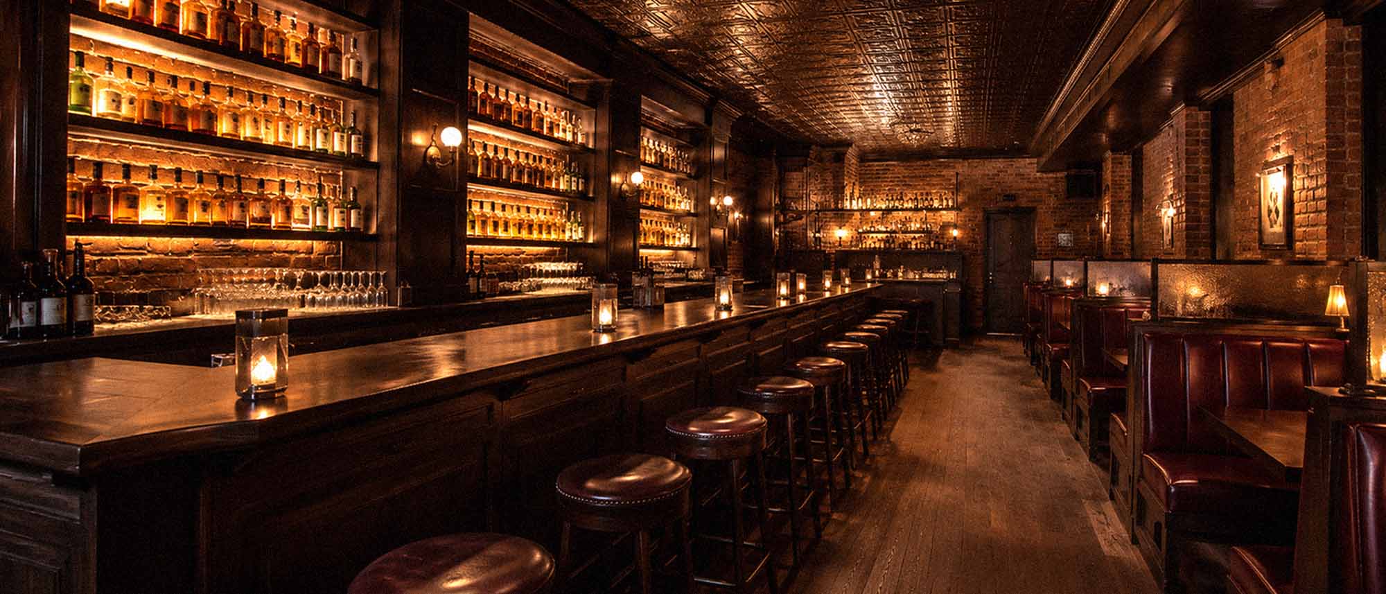 Dark and cozy bar interior with wooden bar, stools, and shelves filled with bottles.