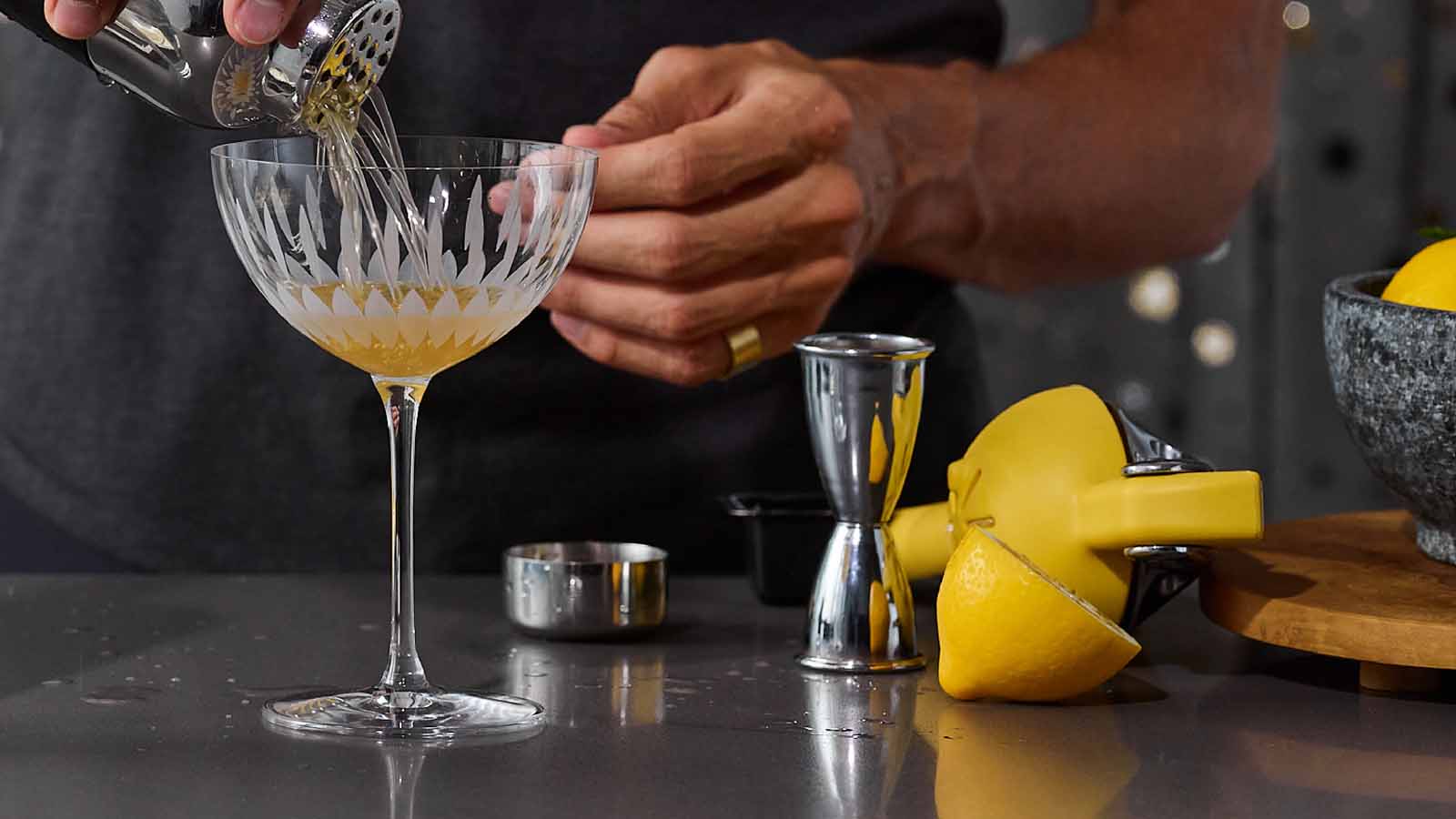 Person pouring a cocktail into a glass with lemon slices and bar tools on a counter.