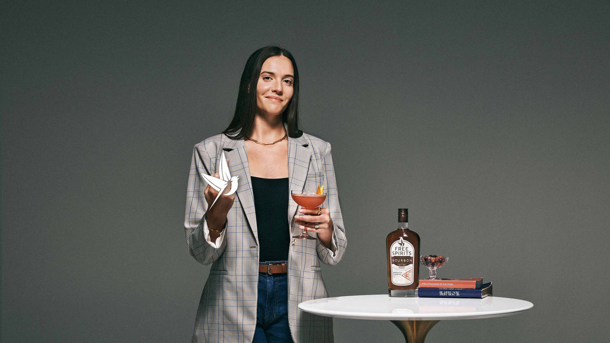 A woman holding a Paper Plane Cocktail with a bottle of Free Spirits