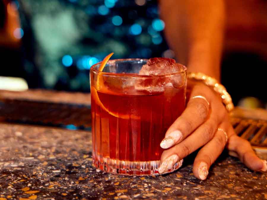 Red cocktail in a glass with ice and an orange peel, held by a hand on a bar counter.