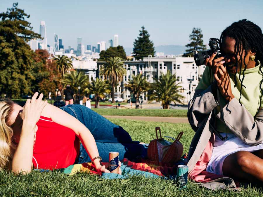 Two people in a park with one taking a photo of the other, city skyline in the background.